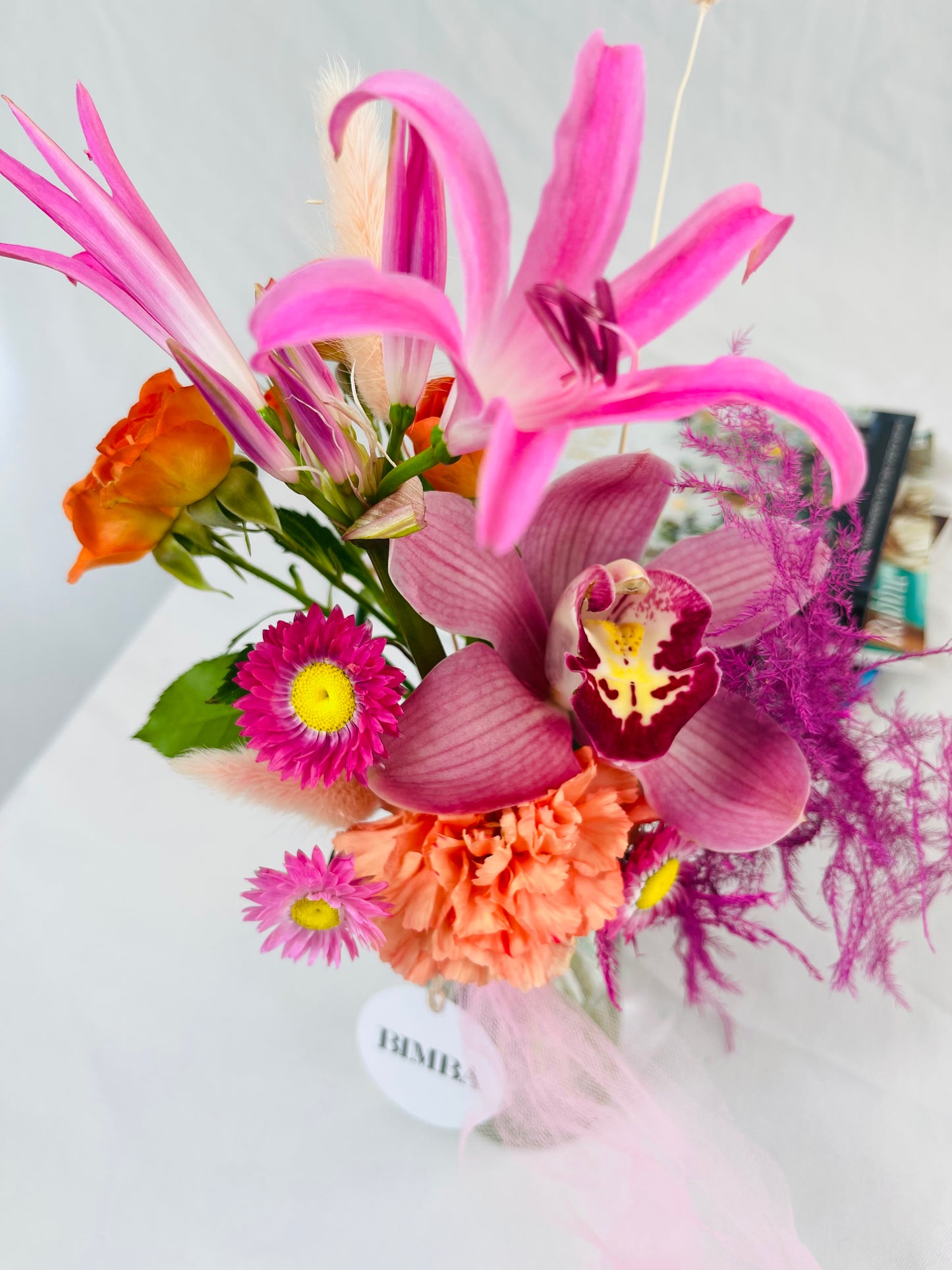 Full of grace mini floral arrangement, in hues of fuchsia and orange tones featuring luxury orchids, eye-catching nerine or french Clematis, sweet scented  mini blooms, timeless carnation, dried conserved acroclinium or camomille flower, asparagus fern and bunny tails.